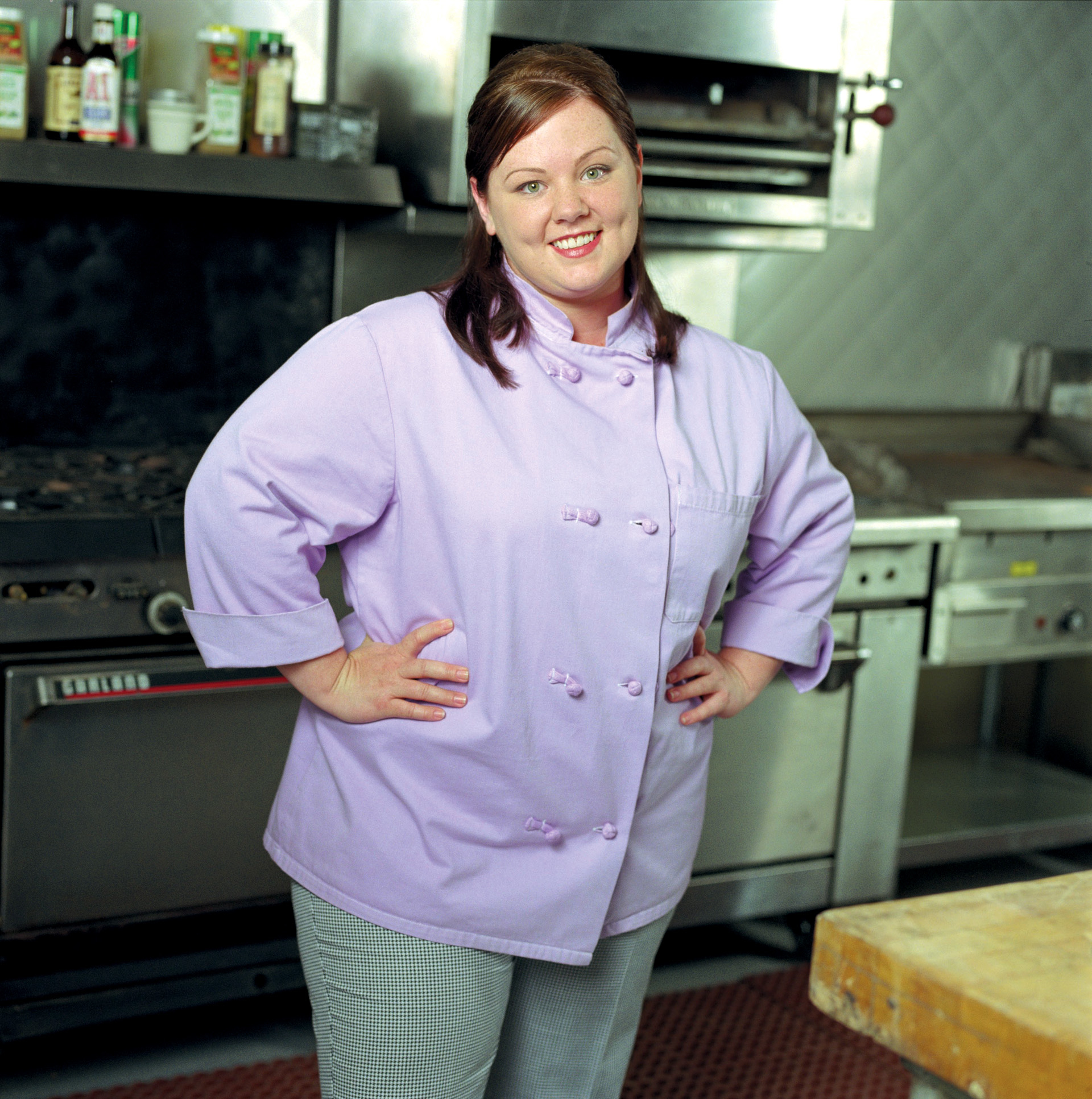 Sookiein a chef's kitchen posing with a smile. She is wearing a traditional double-breasted chef's coat and checked pants