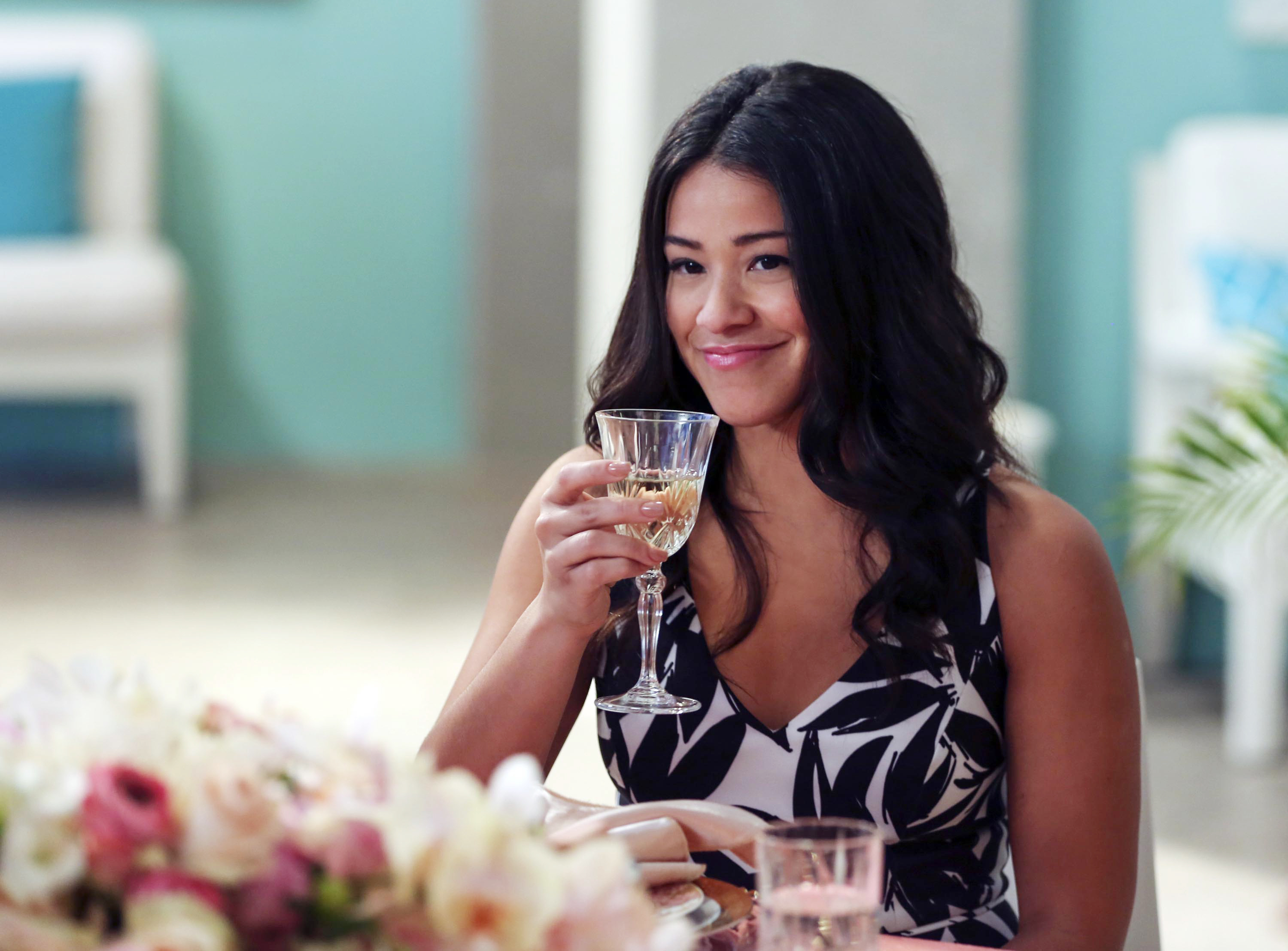 Gina Rodriguez in a printed dress raises a wine glass with a smile while seated at a table with floral arrangements