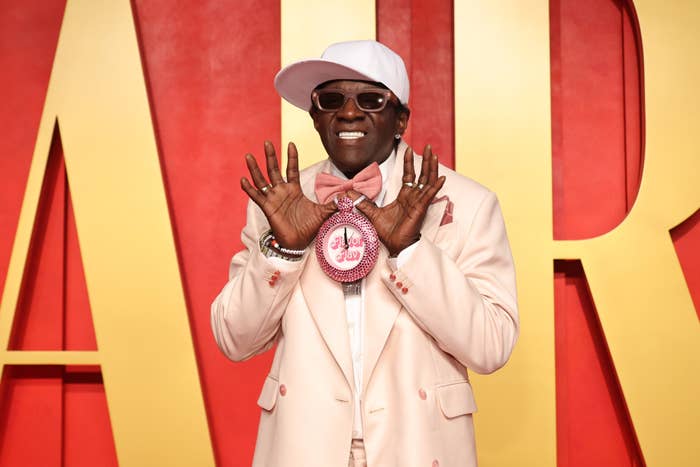 Flavor Flav on red carpet in stylish light-collared suit, bow tie, a fitted hat, and large clock necklace, posing with hands up and smiling