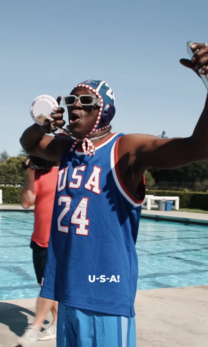 Flavor Flav, wearing a blue &quot;USA 24&quot; jersey and swim cap, enthusiastically chants into a megaphone by a pool