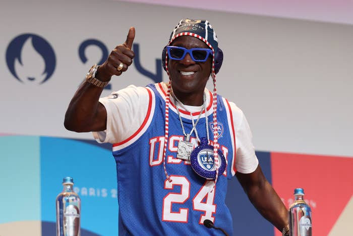 Flavor Flav gives a thumbs up at a sporting event, wearing a blue and white USA basketball jersey and large clock necklace