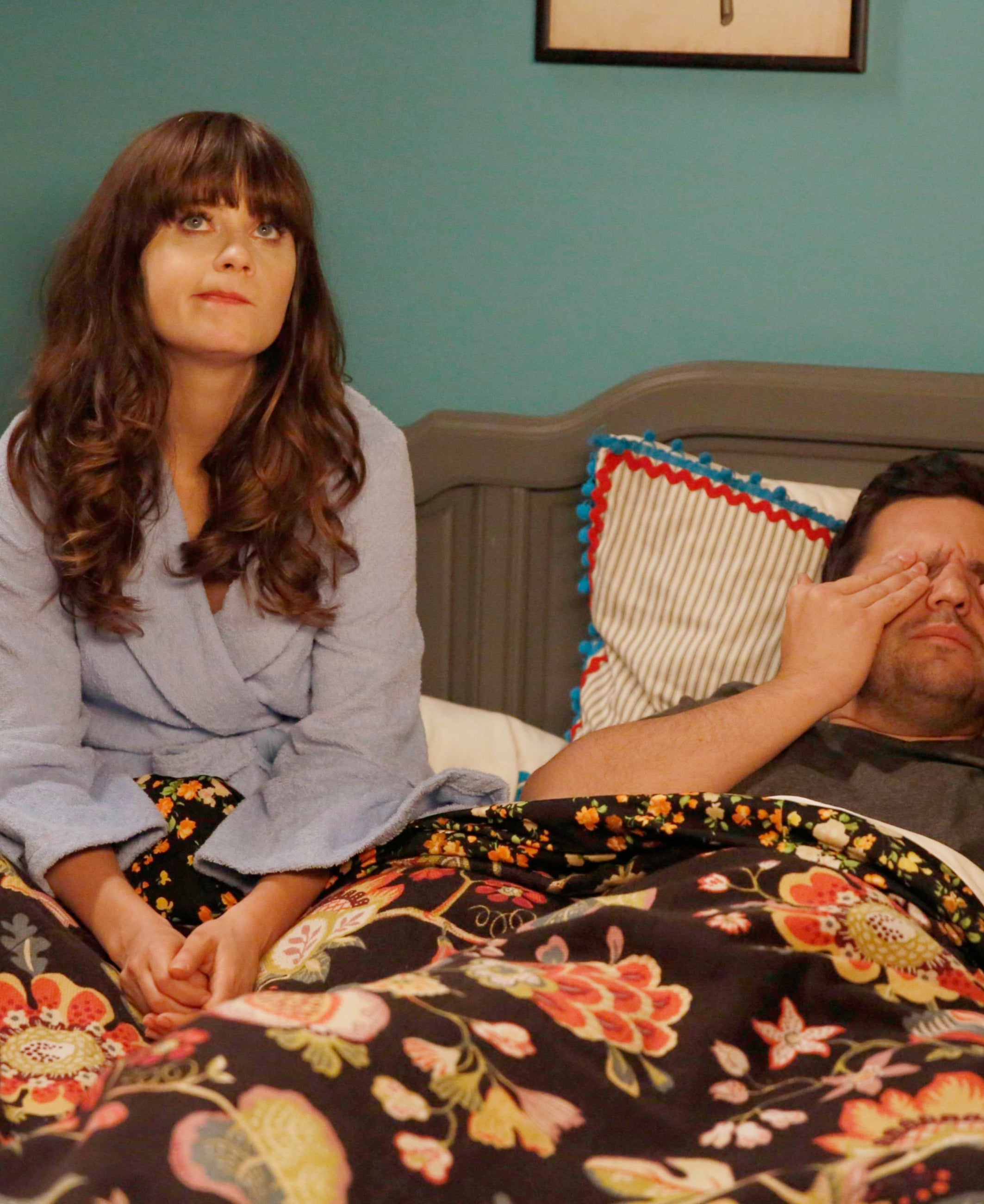 Zooey Deschanel sits on a bed while making a quirky face
