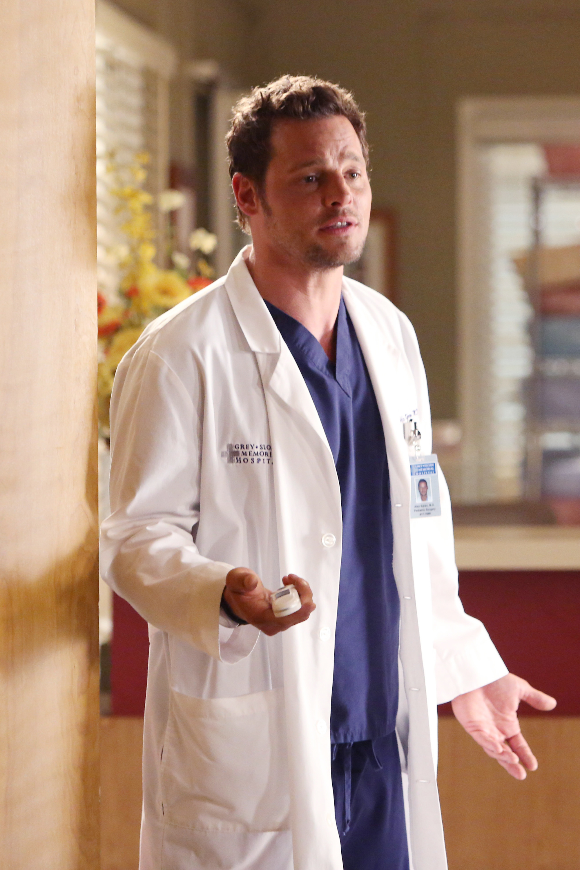 Justin Chambers portraying Dr. Alex Karev in a hospital setting, wearing a lab coat over medical scrubs, holding a pager and gesturing with his other hand