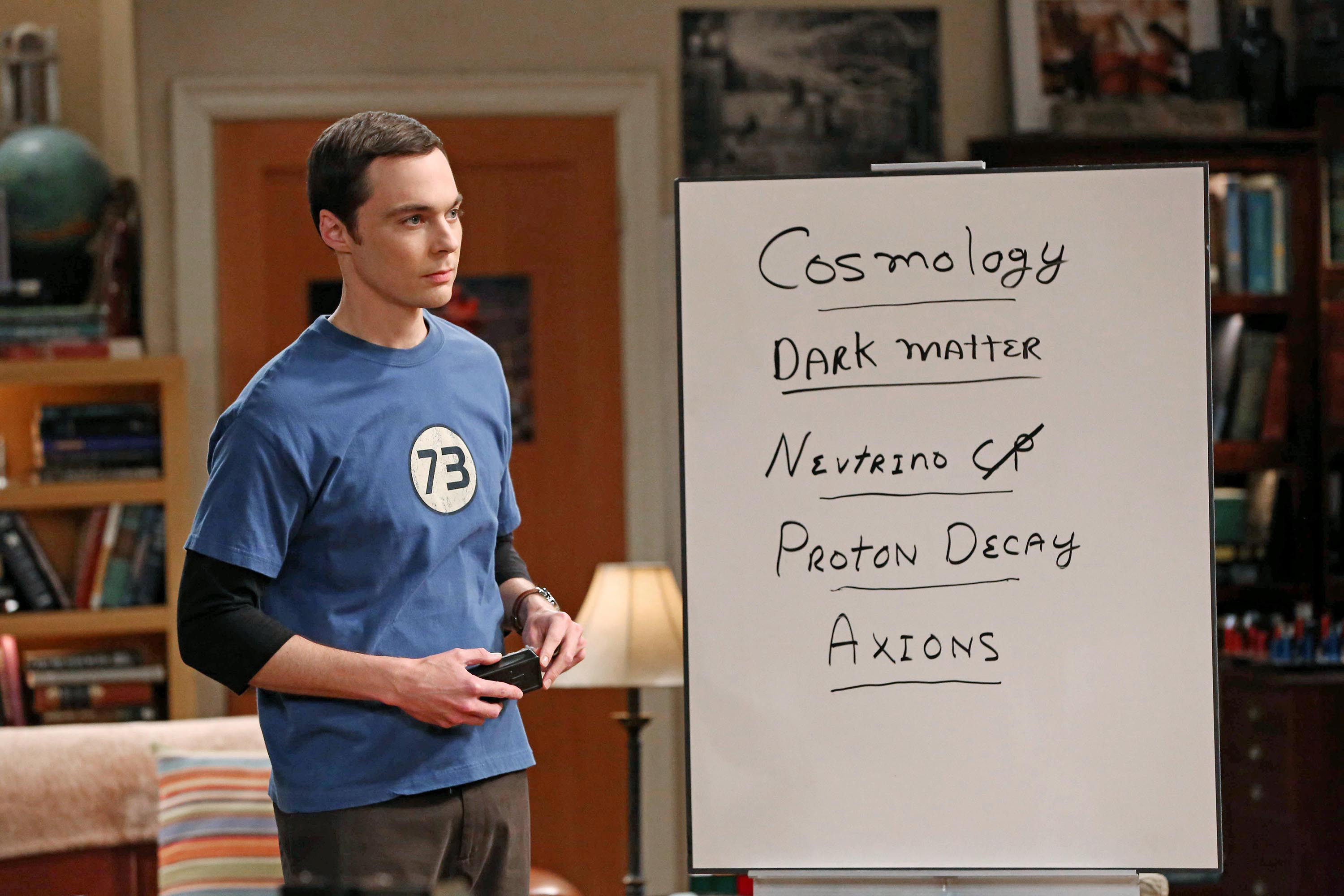 Sheldon Cooper stands next to a whiteboard listing cosmology, dark matter, neutrinos, proton decay, and axions