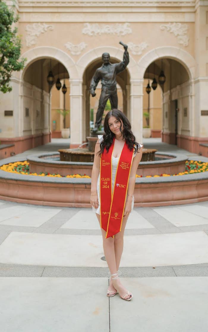 Caitlin stands in front of an arched building and statue wearing a graduation cap and sash that reads "Class of 2024" and "UNC."