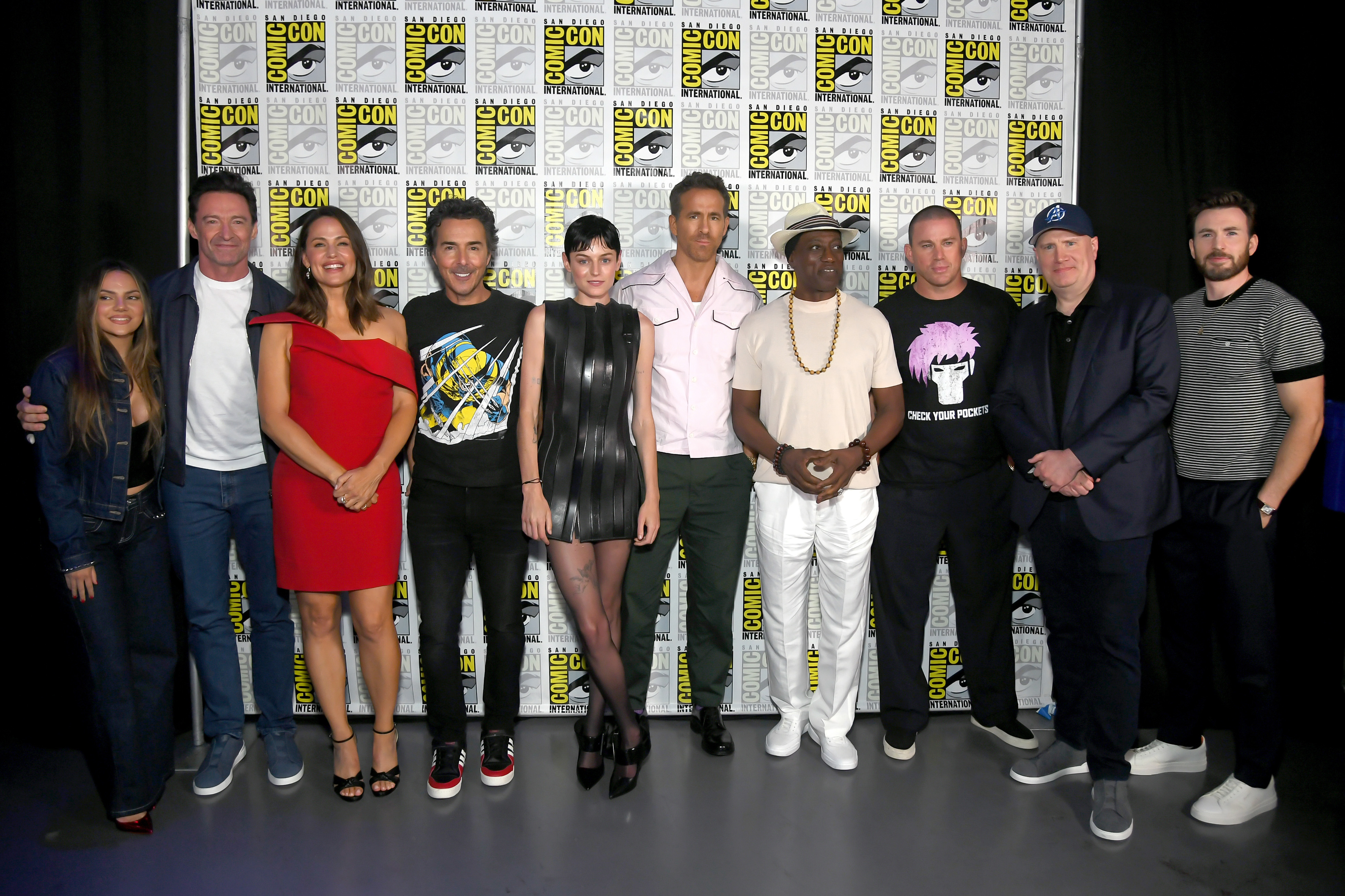 A group photo at Comic-Con featuring stars including Ryan Reynolds, Hugh Jackman, and others standing in front of a banner with Comic-Con logos