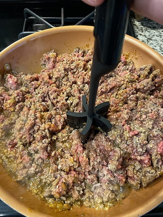 Ground beef being cooked in a skillet, with a hand using a meat chopper tool to break up the meat