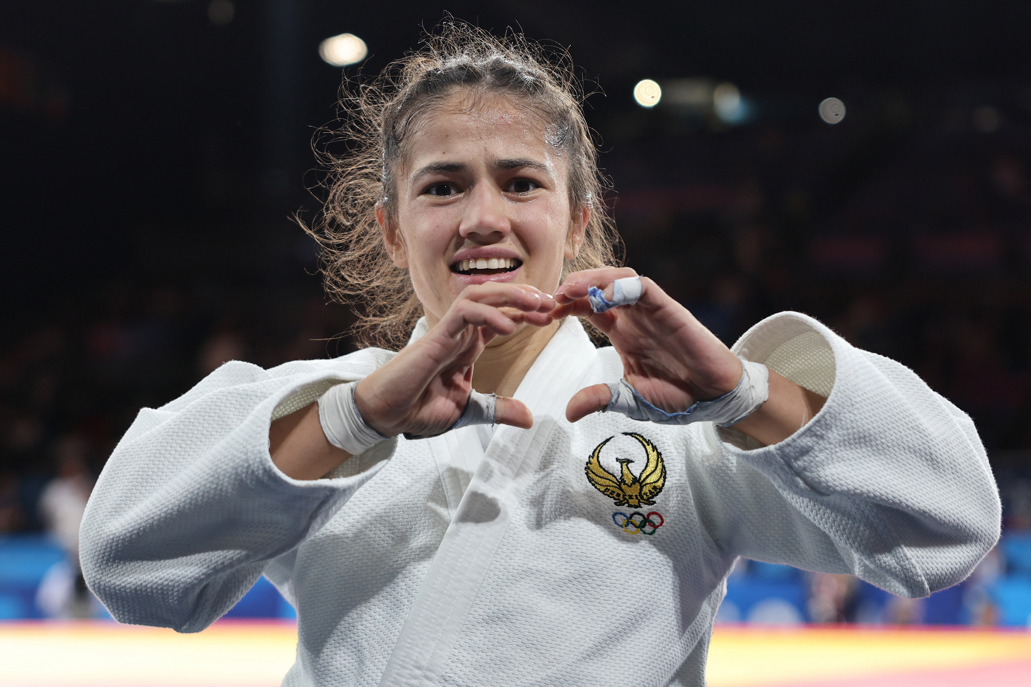 A female athlete wearing a judo gi with Olympic rings is smiling and making a heart shape with her hands