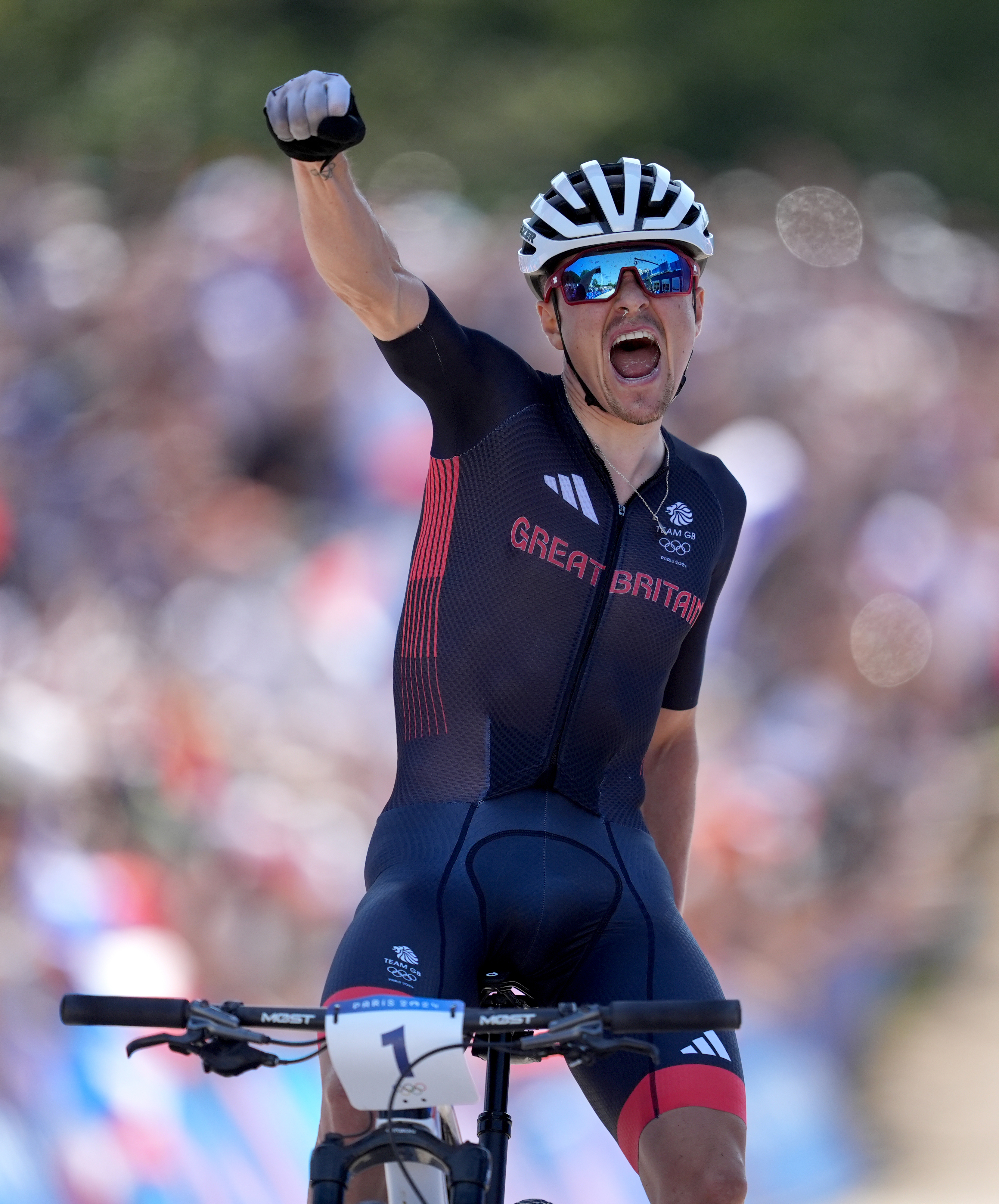 Mountain biker wearing a Great Britain uniform and helmet raises a triumphant fist while crossing the finish line