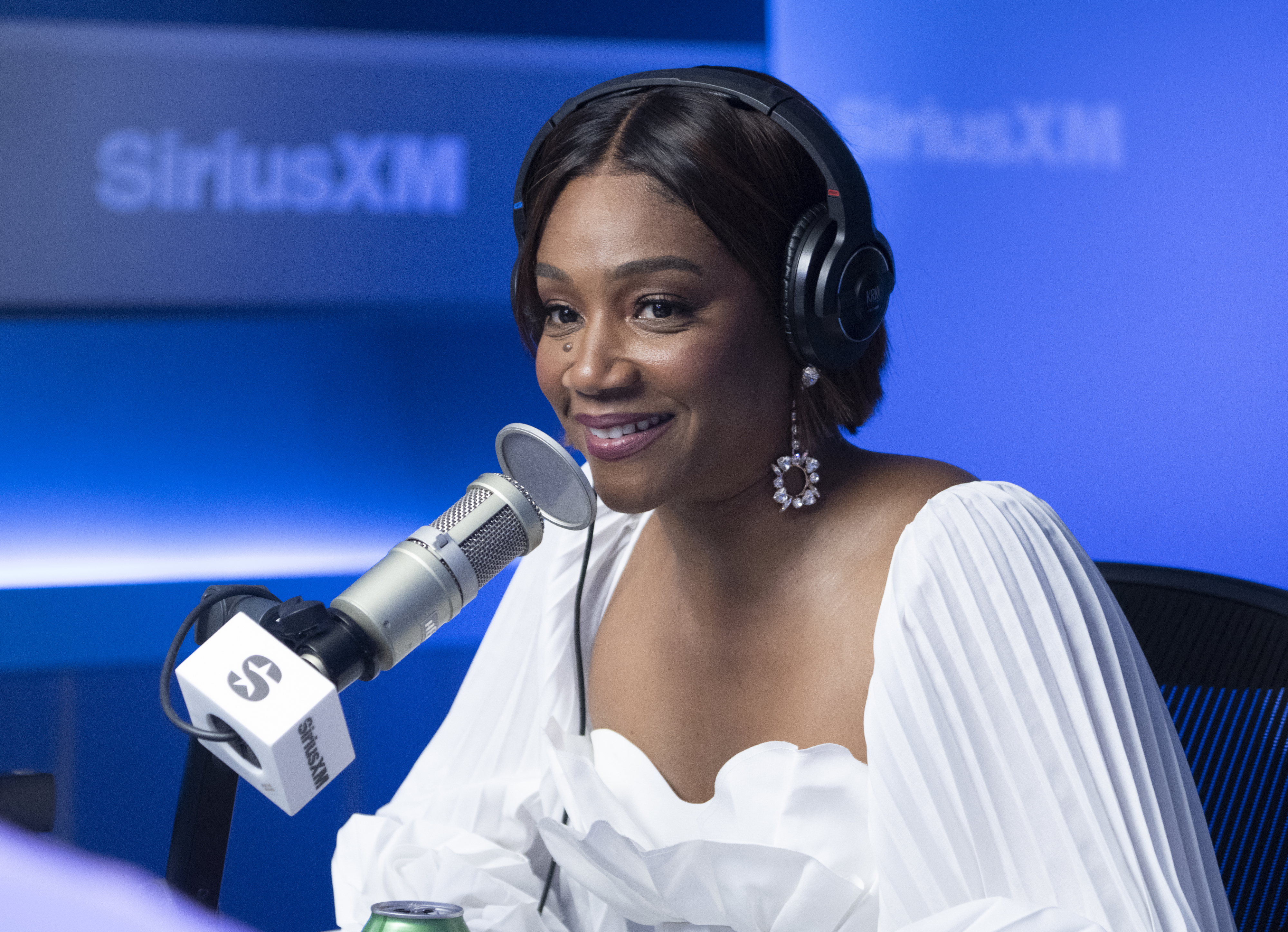 Tiffany Haddish speaks into a microphone during a SiriusXM radio interview, wearing headphones, a white off-shoulder blouse, and dangling earrings