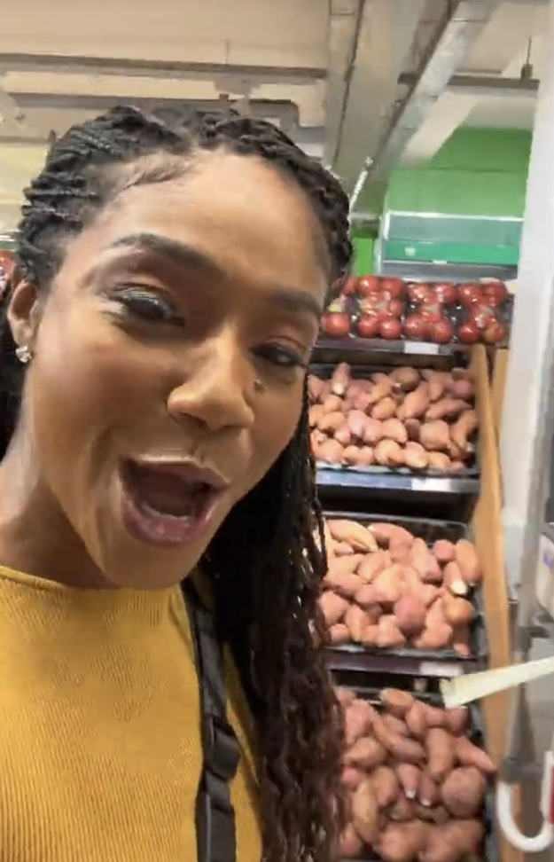 A woman with braided hair, in a grocery store, smiles while standing next to a display of sweet potatoes