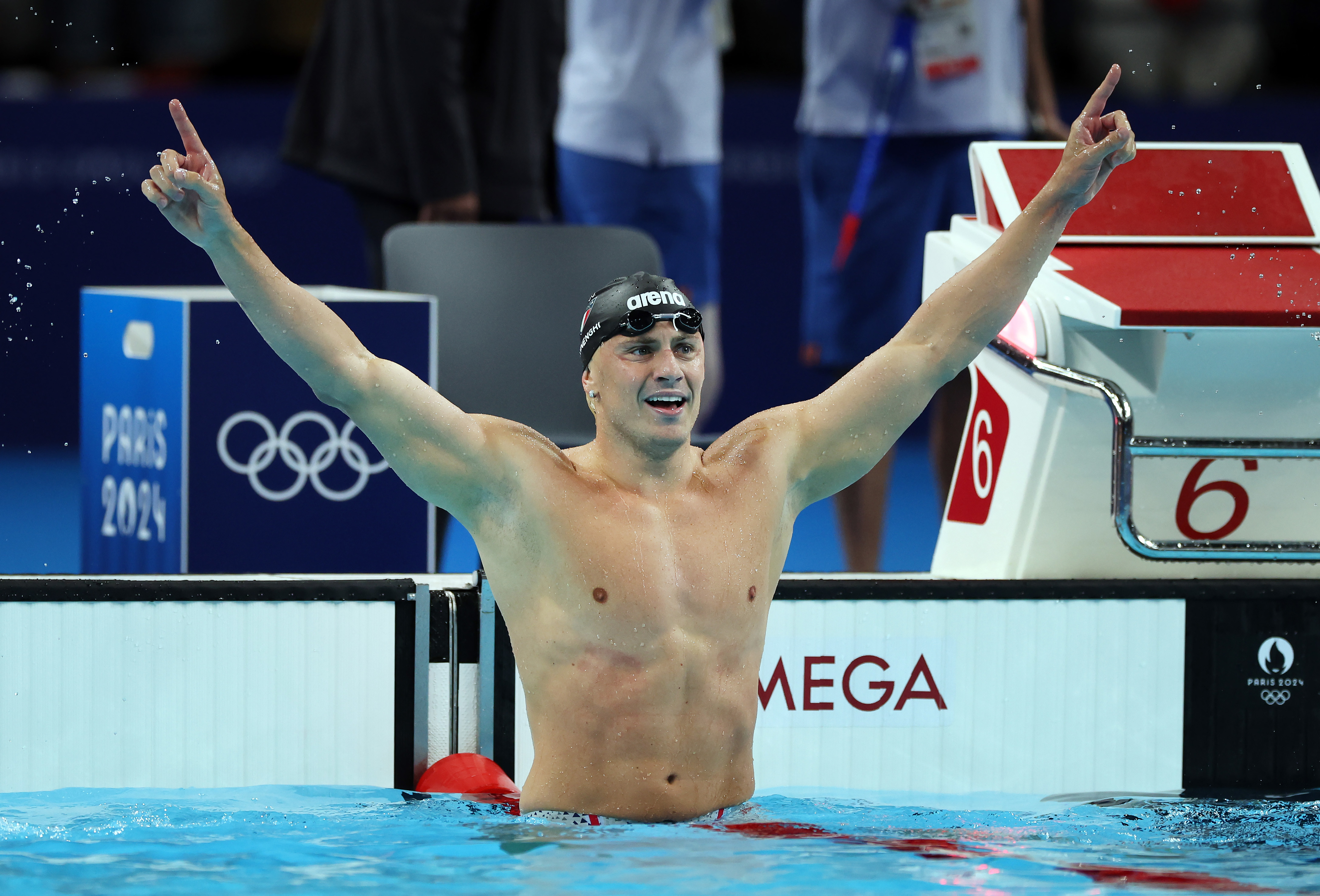 A swimmer, wearing a swim cap and goggles, celebrates with arms raised in a pool next to a 'Paris 2024' sign