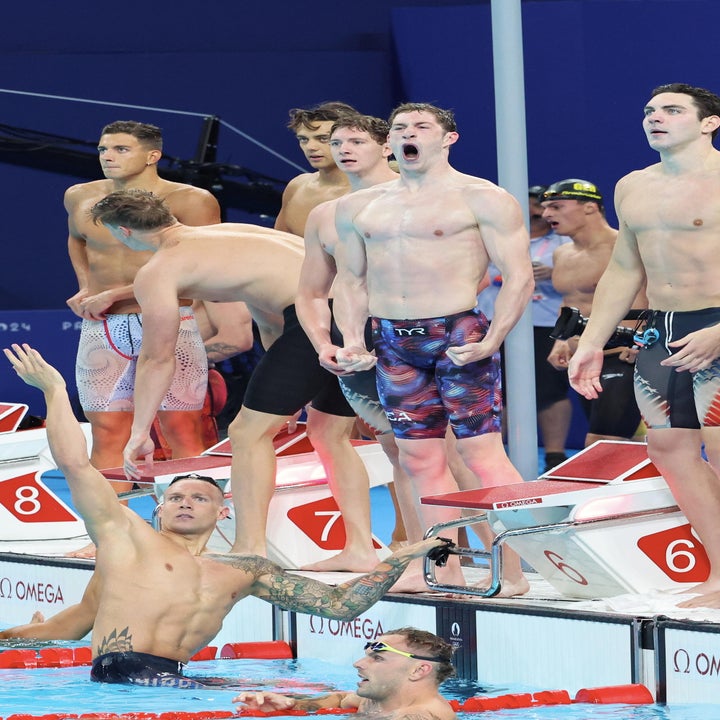 A group of male swimmers, including one pointing upwards, react energetically beside a pool during a swimming competition