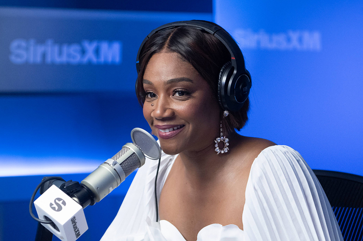 Tiffany Haddish, wearing a white off-shoulder dress and large earrings, speaks into a microphone during a SiriusXM interview