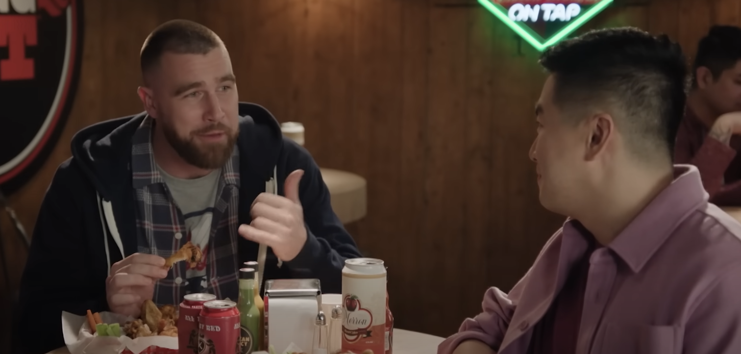 Travis Kelce, wearing a plaid shirt and jacket, enjoys wings and converses with Bowen Yang, who's dressed in a casual button-up at a bar table with drinks and condiments