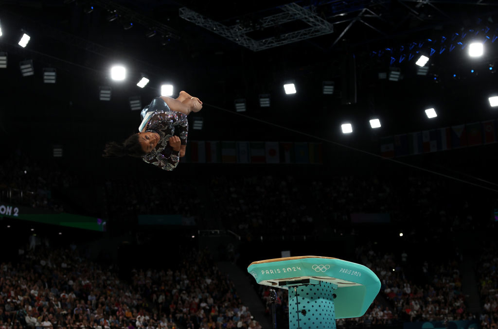 A gymnast performs a mid-air flip over a vault during the Paris 2024 Olympics, with a large audience and bright lights in the background