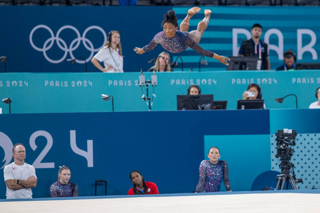 Simone Biles performs a floor routine at the 2024 Paris Olympics. Several officials and gymnasts, including Suni Lee, watch in the background