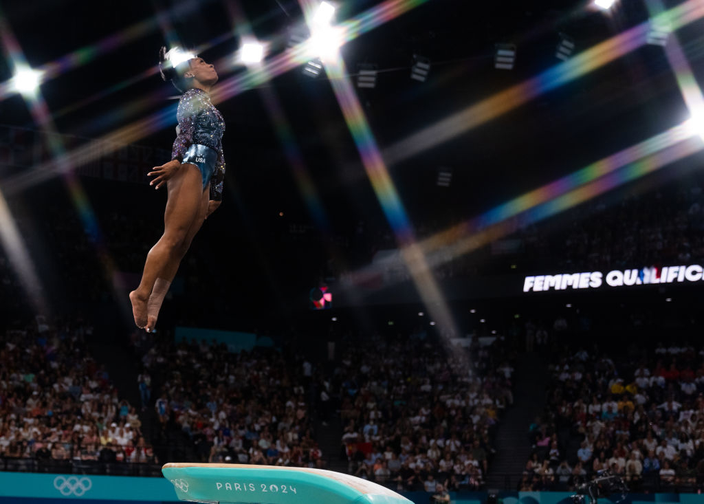 A gymnast performs a high leap on the balance beam at an Olympic event. A large audience watches in the background. "Paris 2024" is visible on the beam