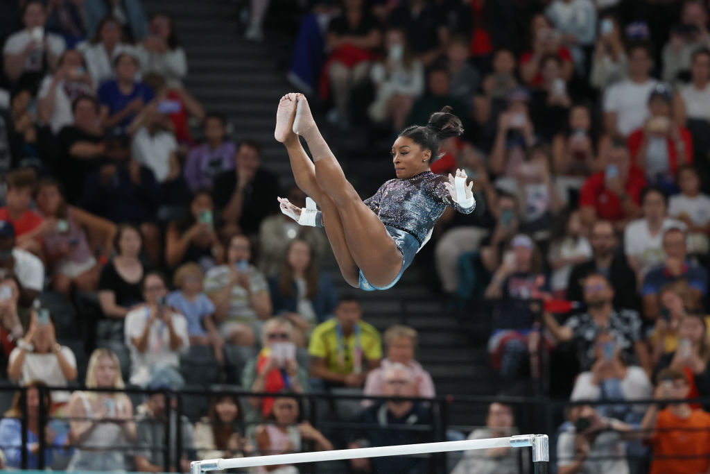 Simone Biles performs a skill above the uneven bars during a gymnastics competition, with a crowd in the background