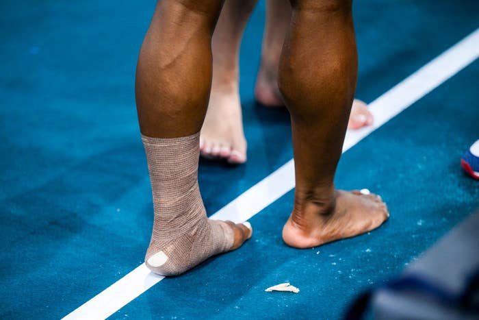 Close-up of an athlete's legs and feet on a gym floor, with one ankle wrapped in medical tape