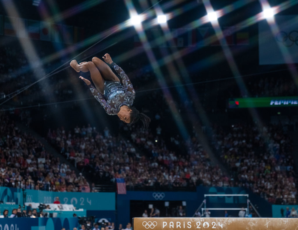 Simone Biles performs a mid-air flip at the Paris 2024 Olympics, dressed in a sparkling gymnastics outfit, in front of a large crowd in an indoor arena