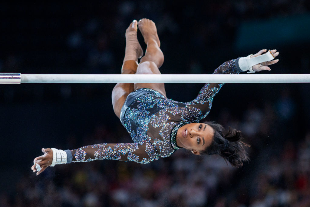 Simone Biles performs a gymnastic routine on the uneven bars, wearing a sparkly leotard with a sheer, long-sleeve design