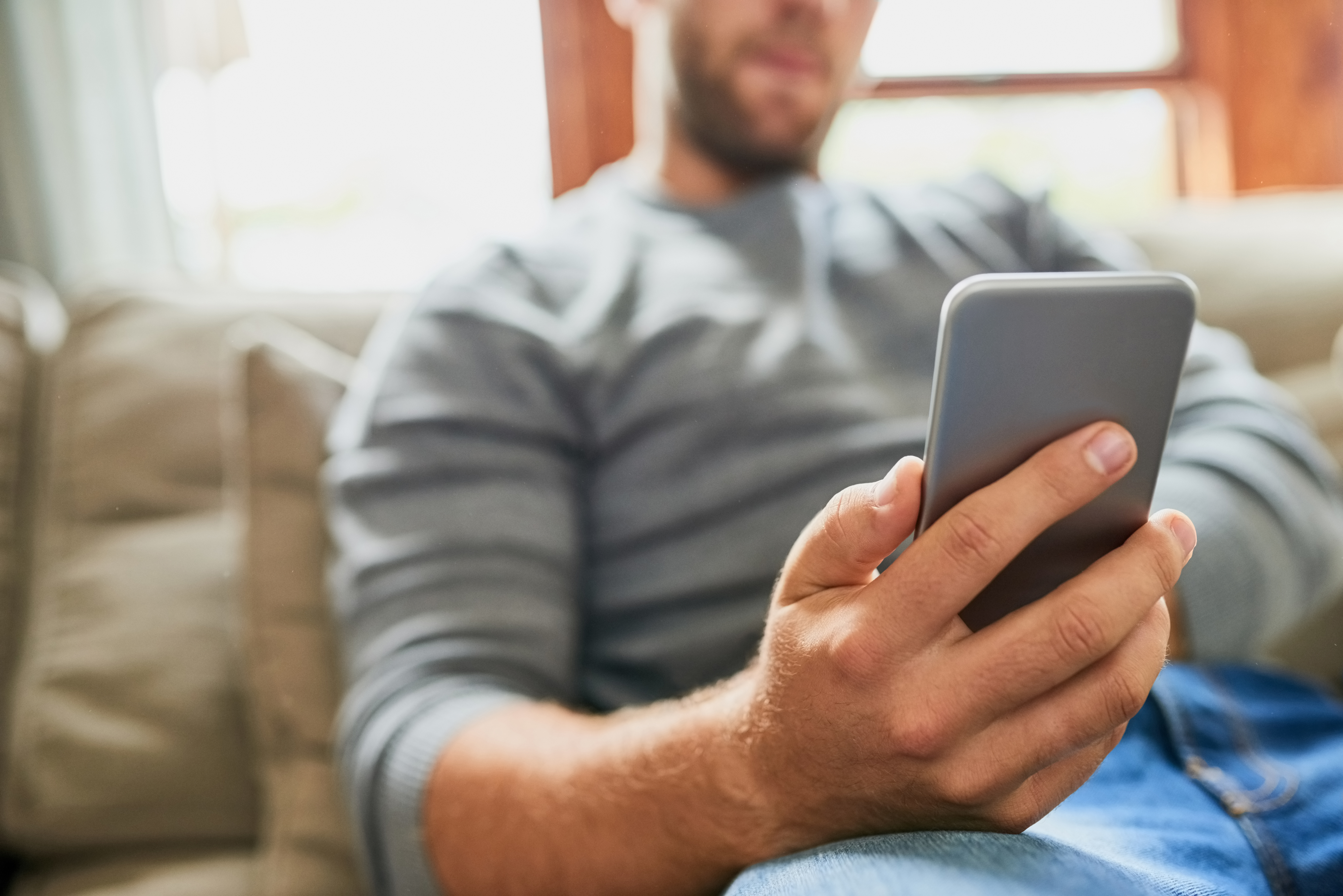 A man, whose name is unknown, is sitting comfortably on a couch, focusing on his smartphone. The image suggests engagement with digital communication or dating apps
