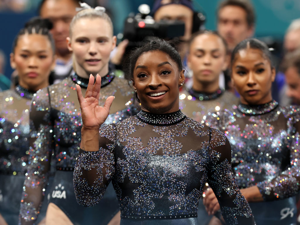 Simone Biles waves while standing with her teammates. They are dressed in sequined gymnastics leotards during a competition