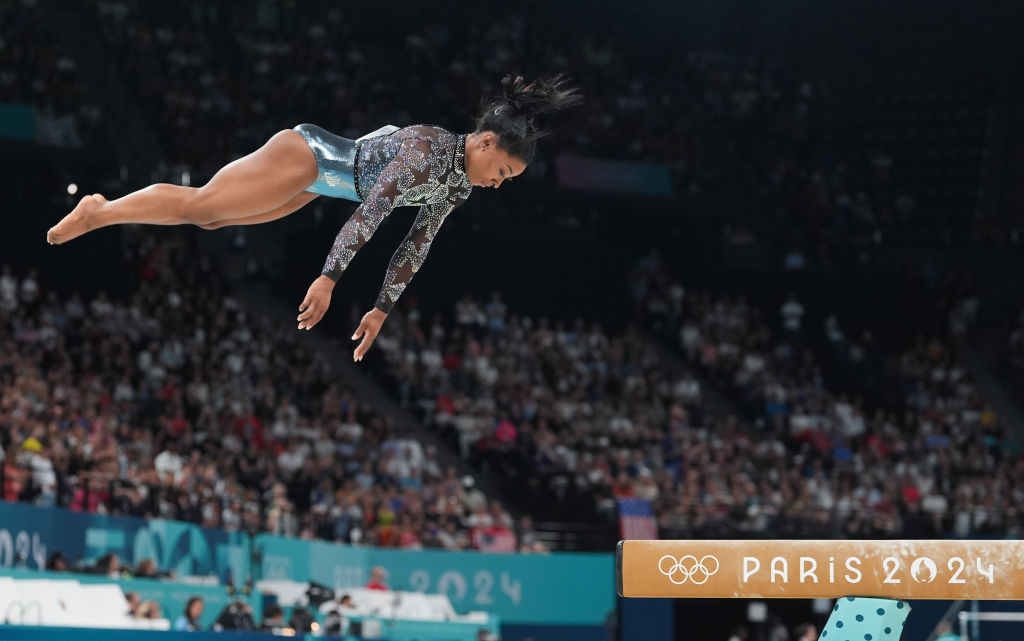 Gymnast Simone Biles performing a jump above a balance beam at the Paris 2024 Olympics, with a focused audience in the background
