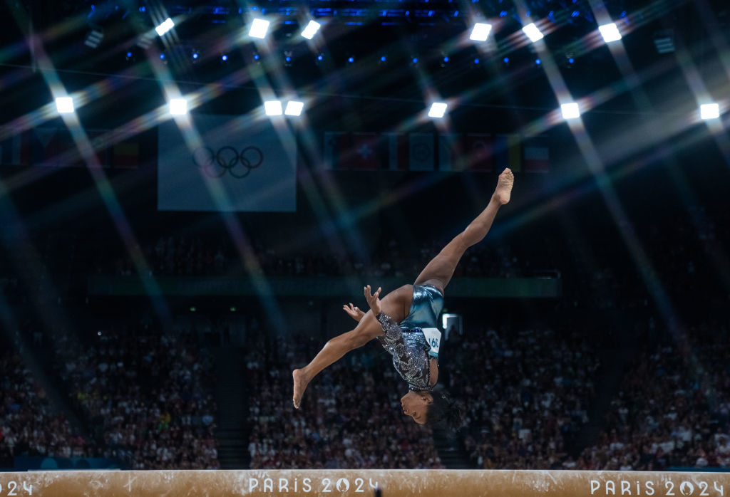 Gymnast in mid-air doing a flip during the balance beam event at the Paris 2024 Olympics, with audience and Olympic rings in the background