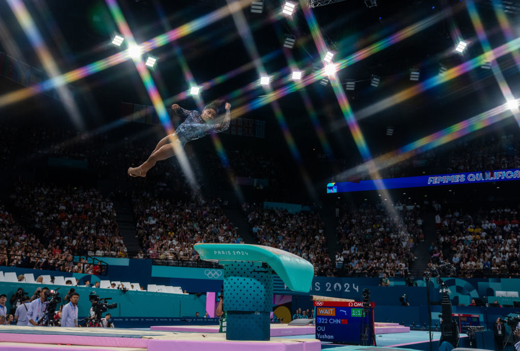 A gymnast mid-air above a vault during an event at the Paris 2024 Olympics in a packed arena. Audience and officials watch in the background