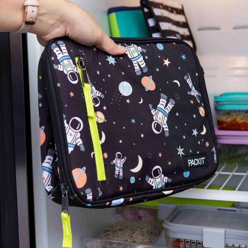 A hand placing a PackIt-brand black lunch bag with astronaut and space-themed design into a refrigerator