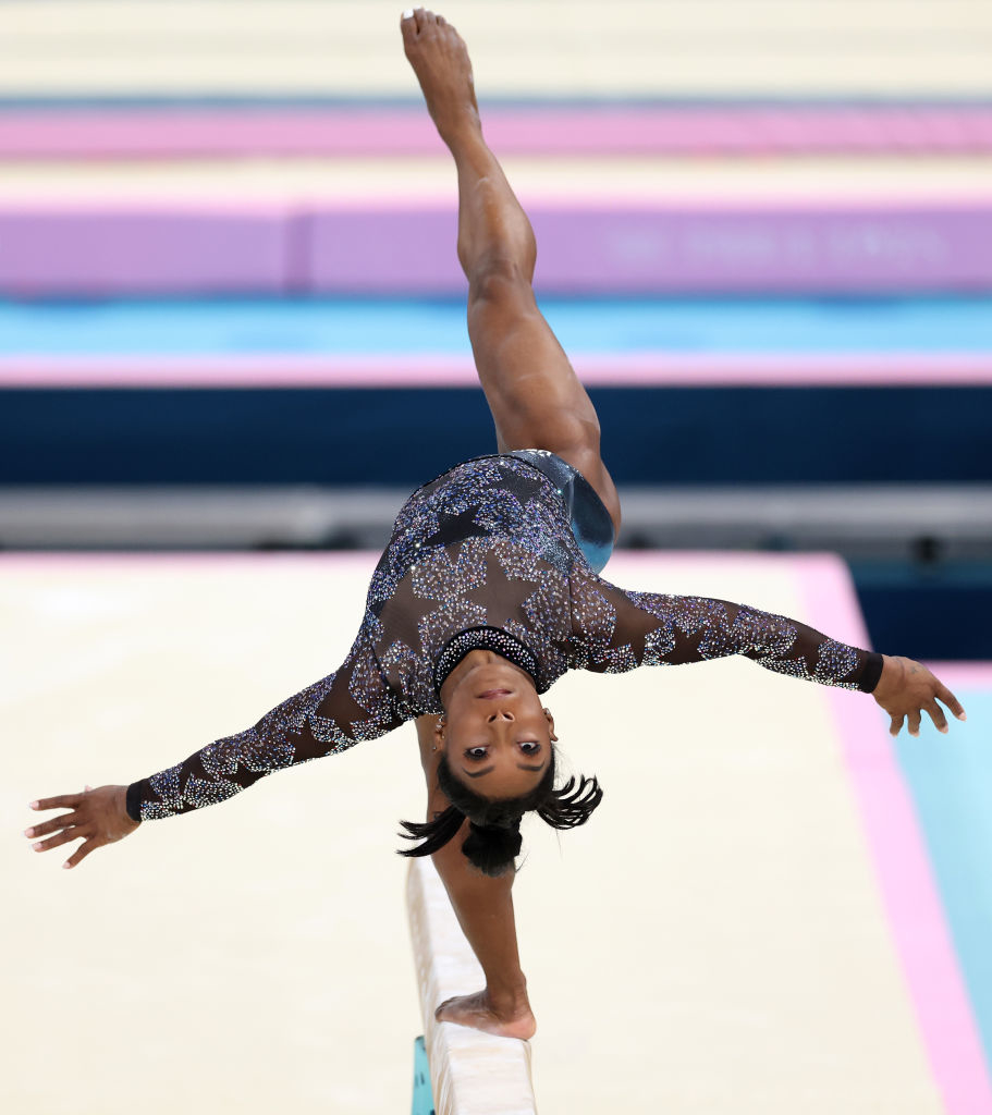 Simone Biles performs an advanced balance beam routine, extending her legs high and arms wide, showcasing her incredible gymnastics skills