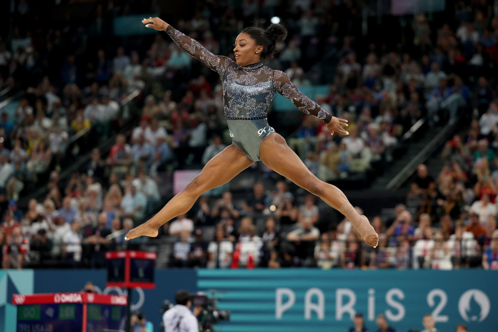 Simone Biles performs a leap in mid-air during a gymnastics routine at a competition in Paris, with an audience in the background