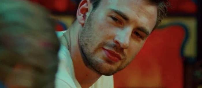 Chris Evans glances over his shoulder with a soft expression, wearing a casual white shirt, set against a blurred indoor background