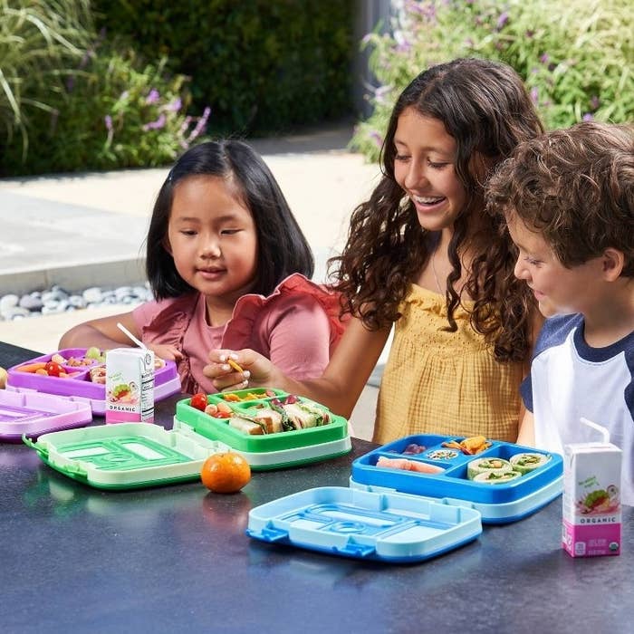 Three children enjoy lunch from colorful lunch boxes in a garden setting
