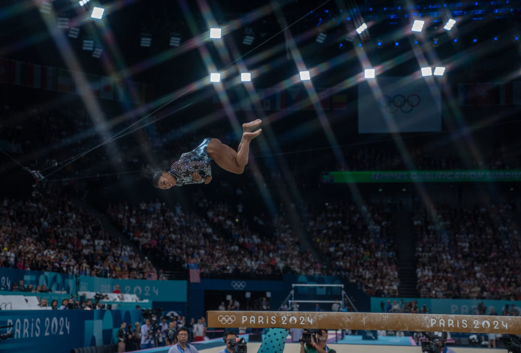 Simone Biles mid-air during a gymnastics performance at the Paris 2024 Olympics, performing a high-difficulty maneuver above the balance beam, with spectators in background