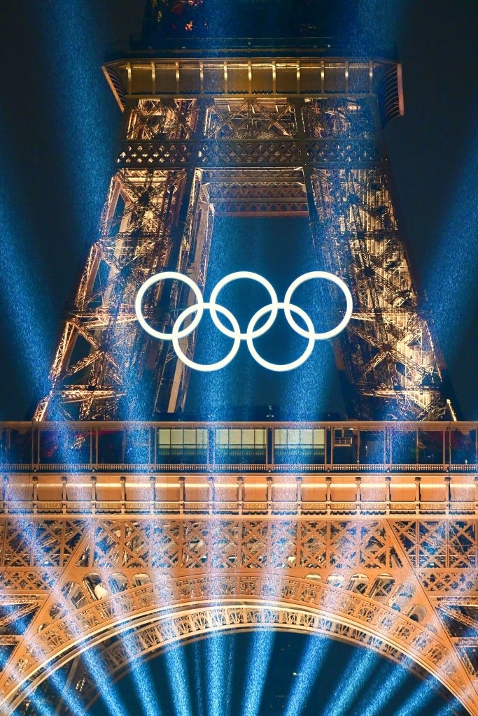 Eiffel Tower lit up at night with the Olympic rings glowing in front, surrounded by bright light beams, signifying the Olympics event