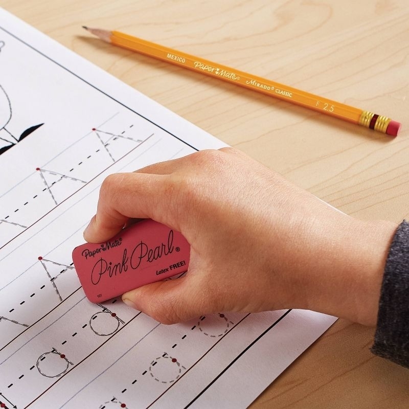 A hand erases handwriting practice lines on paper with a pink Paper Mate Pink Pearl eraser. A Paper Mate pencil is placed on the desk nearby