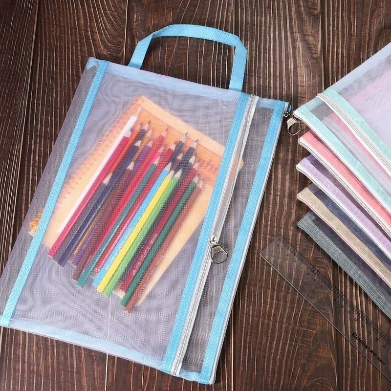 A blue-handled mesh zip bag holds colorful pencils and a notebook, with additional mesh bags and a ruler nearby on a wooden surface
