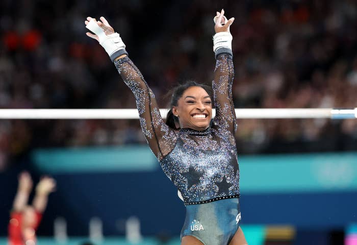 Simone Biles celebrates with raised arms in front of uneven bars, wearing a glittery leotard with "USA" on it