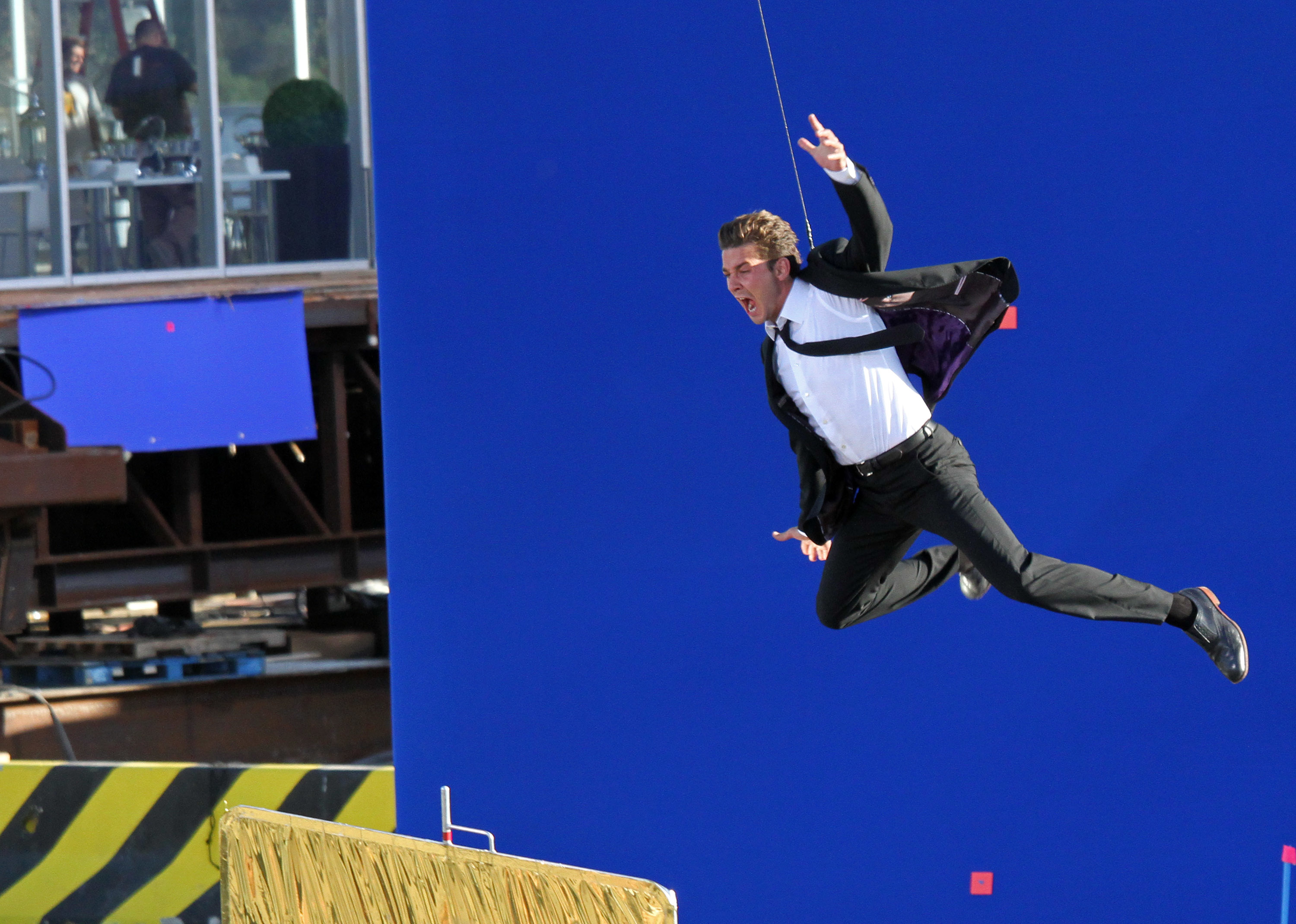 A man in a suit performs a high-flying stunt on a movie set, with visible cables and a blue screen backdrop