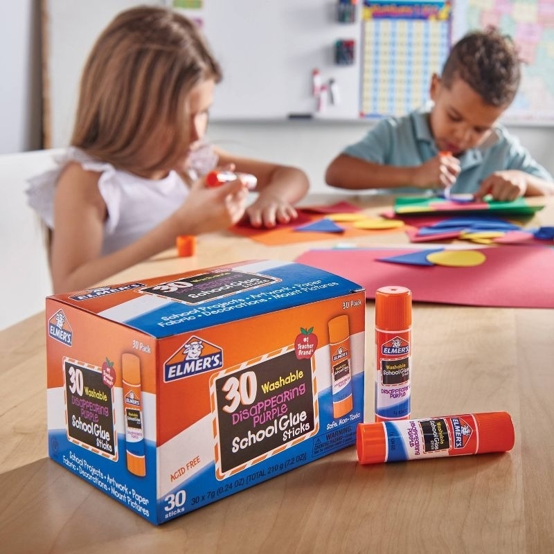 Two children sit at a table crafting with Elmer's Disappearing Purple School Glue Sticks. A box of 30 glue sticks is in the foreground