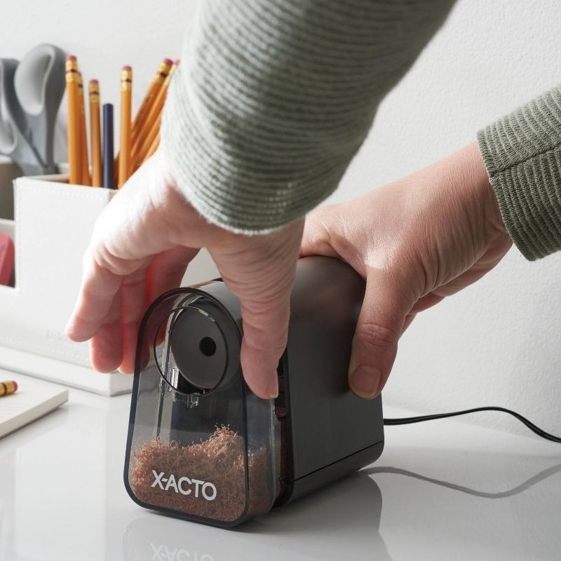 A person inserts a pencil into an X-ACTO electric pencil sharpener on a desk, with other office supplies in the background