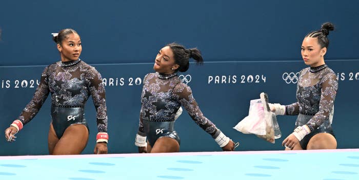 Gabby Douglas, Simone Biles, and Sunisa Lee in black leotards with glittery details, preparing for their routines at the Paris 2024 Olympics