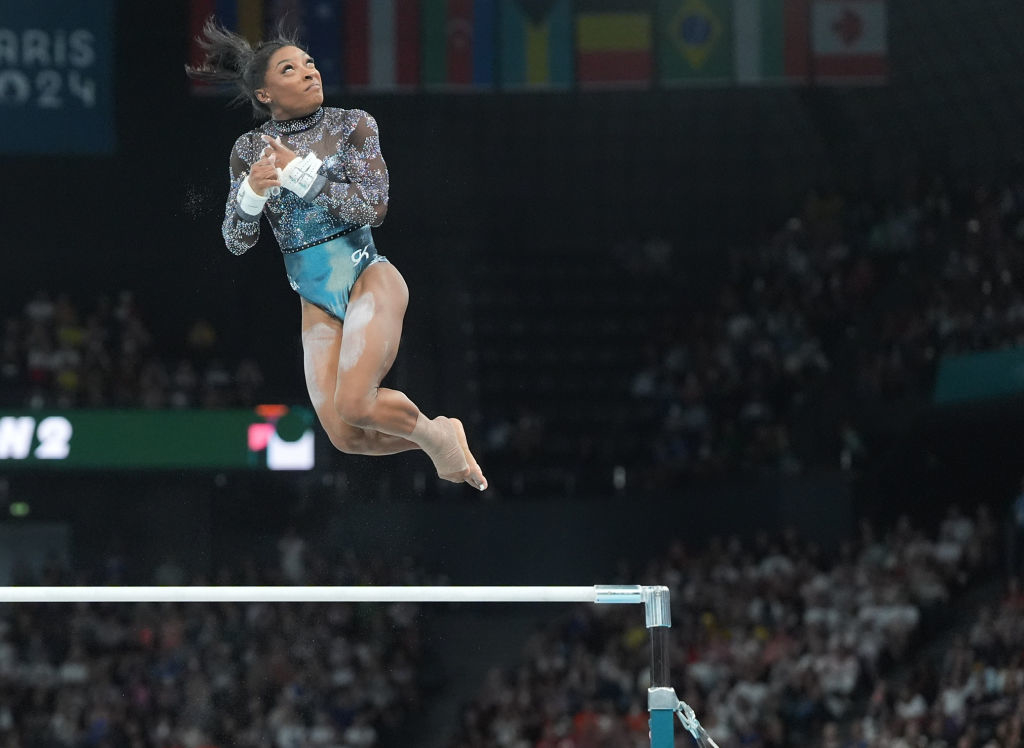 Simone Biles performing on the uneven bars at gymnastics event, mid-air with focused expression, wearing a sequined leotard
