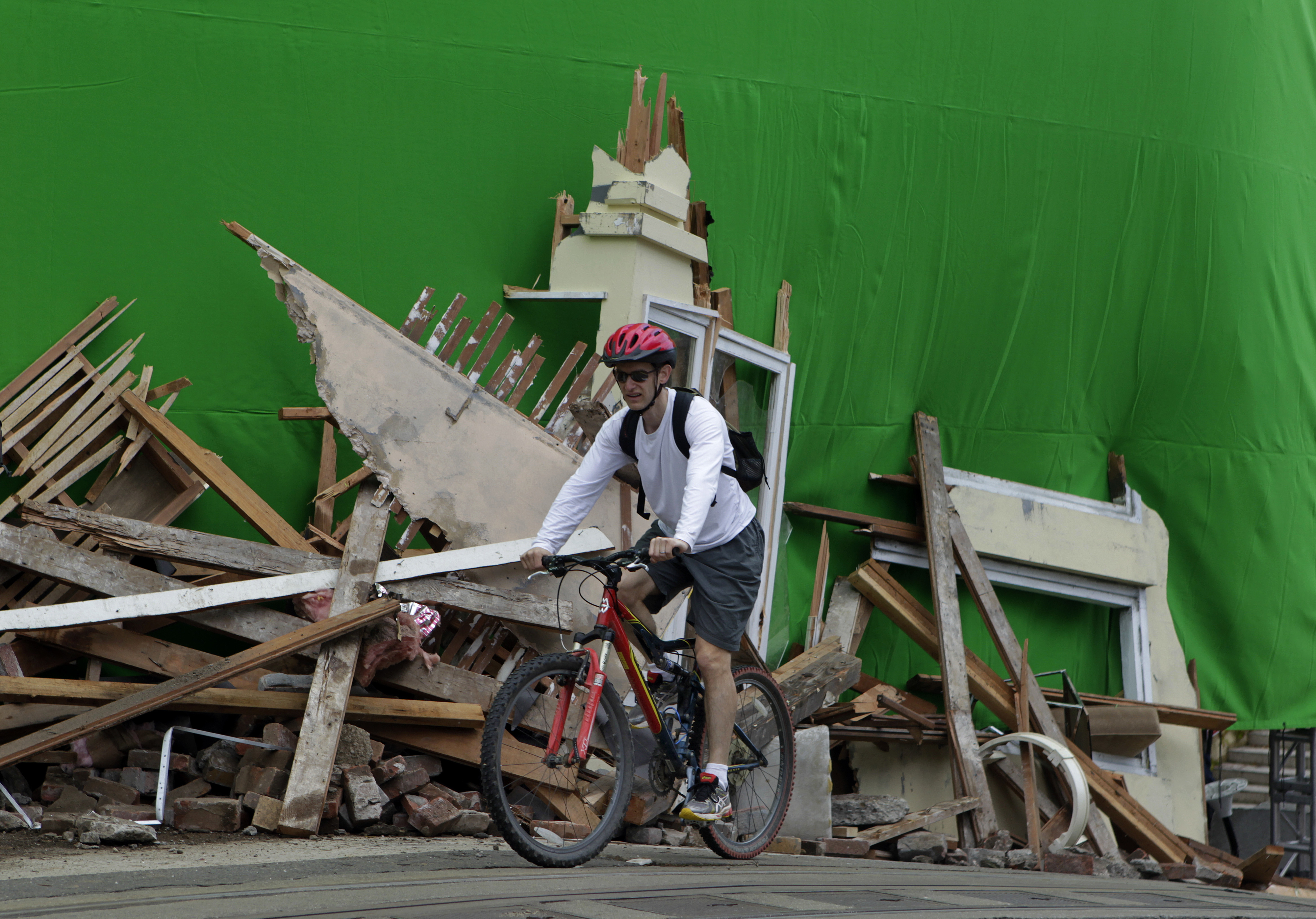 A person rides a bicycle past a large pile of construction debris, with a green screen backdrop behind them