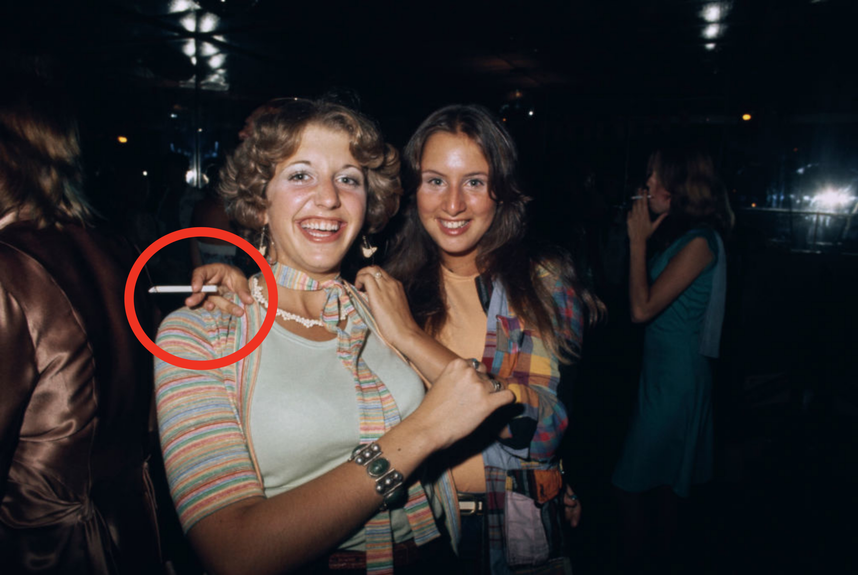 Two women smiling at a party, wearing 1970s outfits with striped patterns, one holds a cigarette