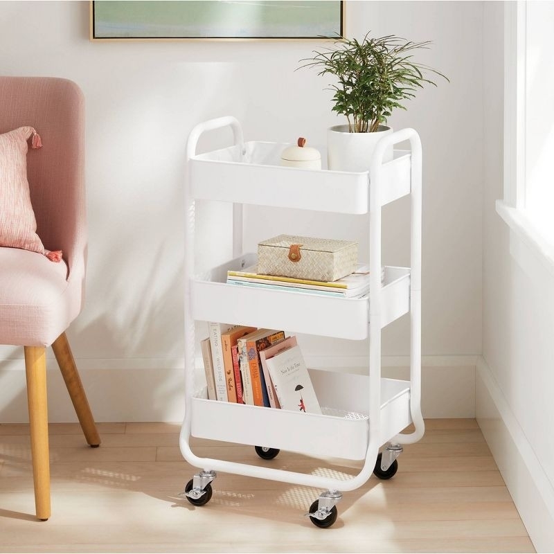 White rolling cart with three shelves, holding a plant, a candle, a small woven box, and various books. Placed next to a pink upholstered chair