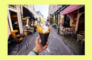 A hand holds a cone of fries with sauce on a cobblestone street, with outdoor seating and shops in the background